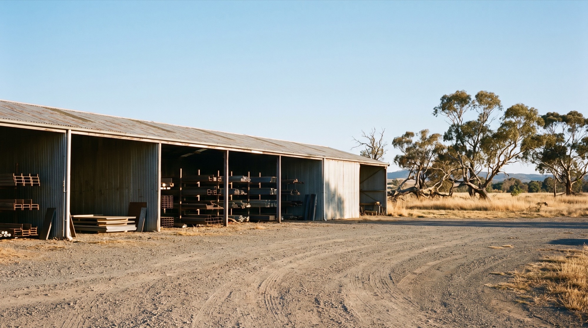 A long corrugated iron supply shed on a gravel forecourt with dry gum trees behind